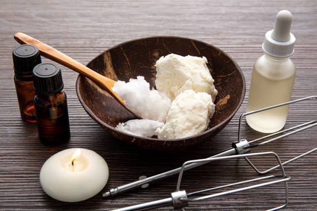 Shea and coconut butter in a coconut bowl with a wooden spoon, amber dropper bottles, a serum bottle, a candle and a whisk